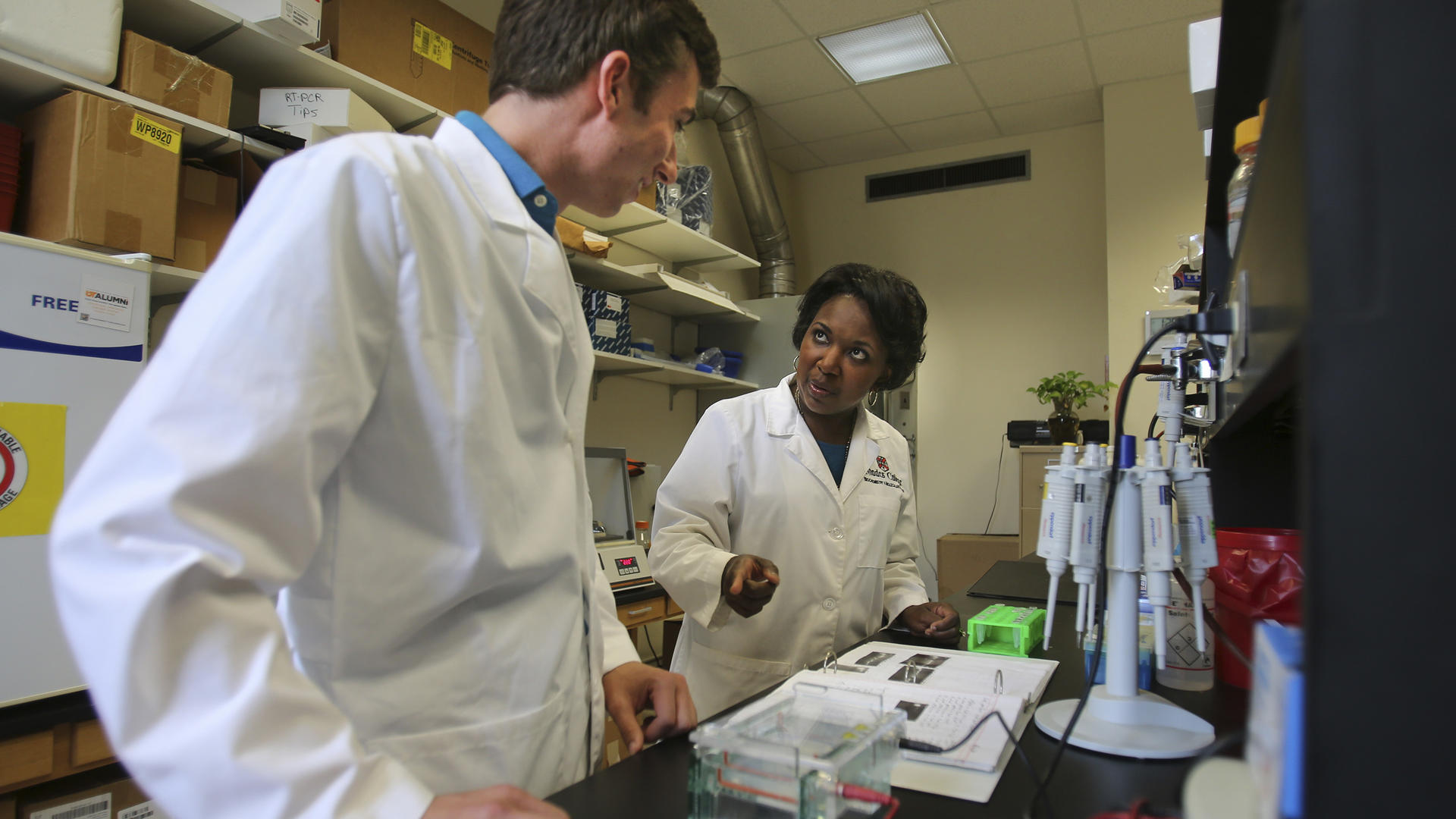 Two researchers in white coats stand at a lab table reviewing notes.