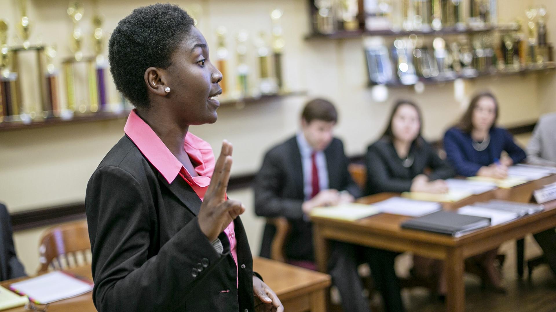 a young woman in a suit speaks before students at a desk in a room lined with trophies