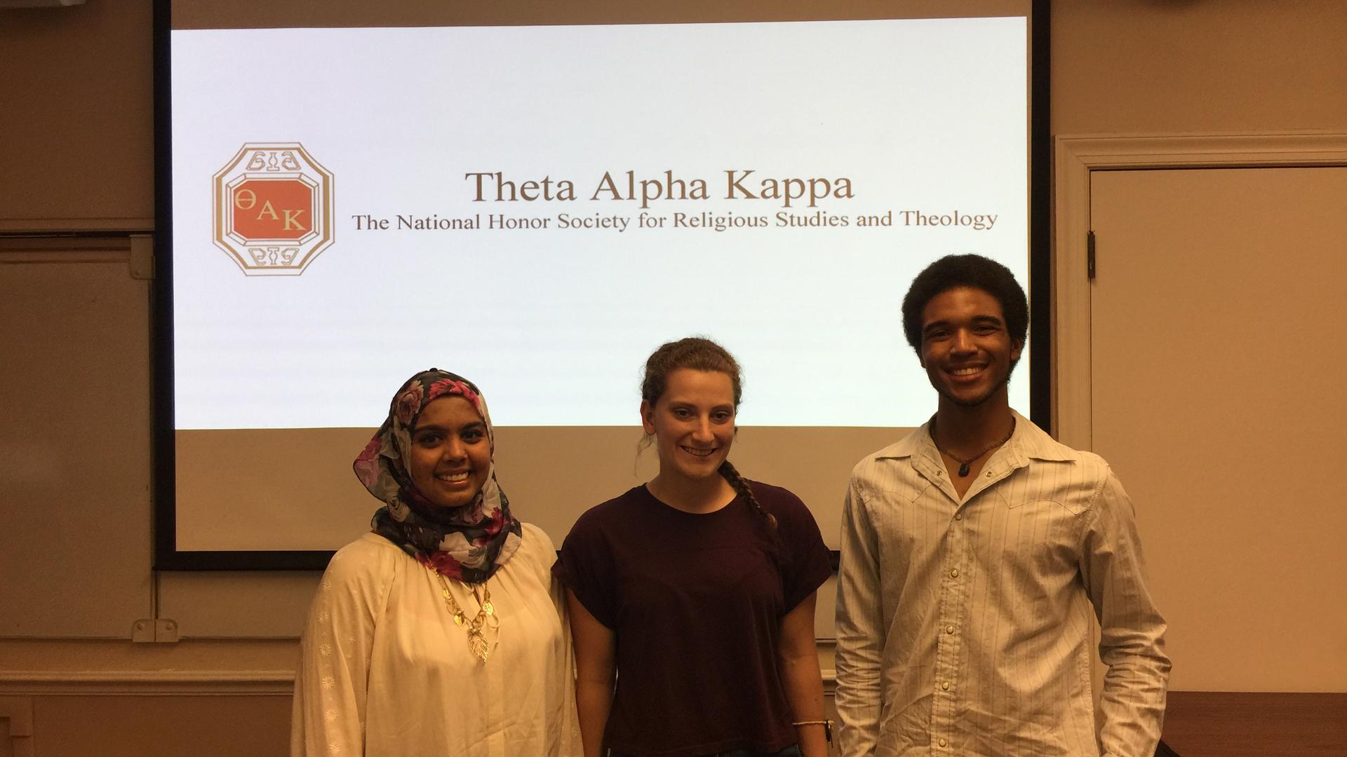Three students stand in front of a projection screen reading Theta Kappa Alpha