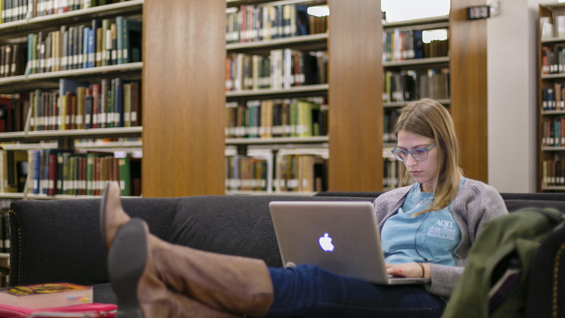 A student sits with her feet up on the coffee table, a laptop on her lap; she is among shelves of books