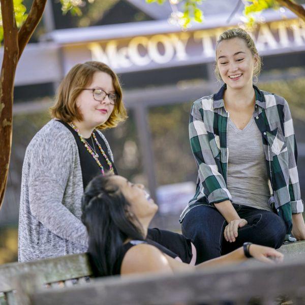 Three students laugh and talk near a bench outside the McCoy Theatre