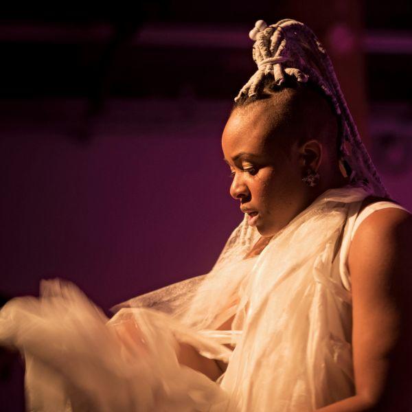 Close up of a black woman wearing flowing white fabric in front of a deep purple backdrop
