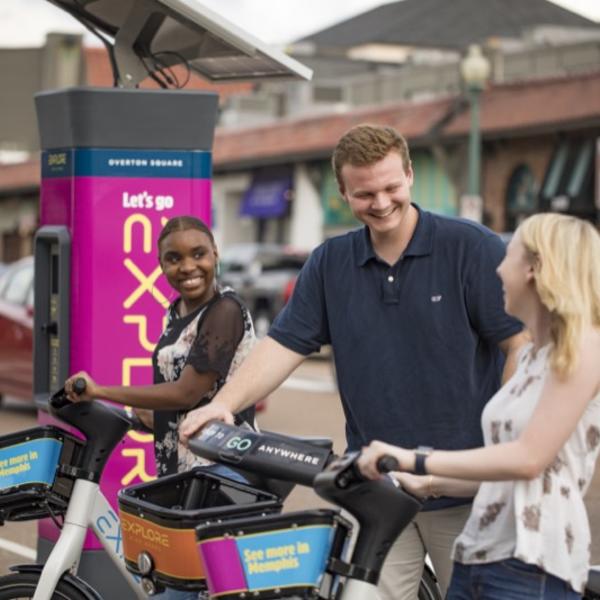 A young man and woman rent bikes from a rideshare station in a small shopping center.