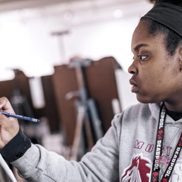 A young woman sketches with a drawing board in an open studio space. 