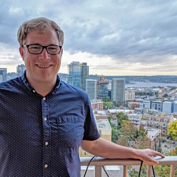 A young man in a blue shirt stands on a balcony overlooking the Seattle skyline.