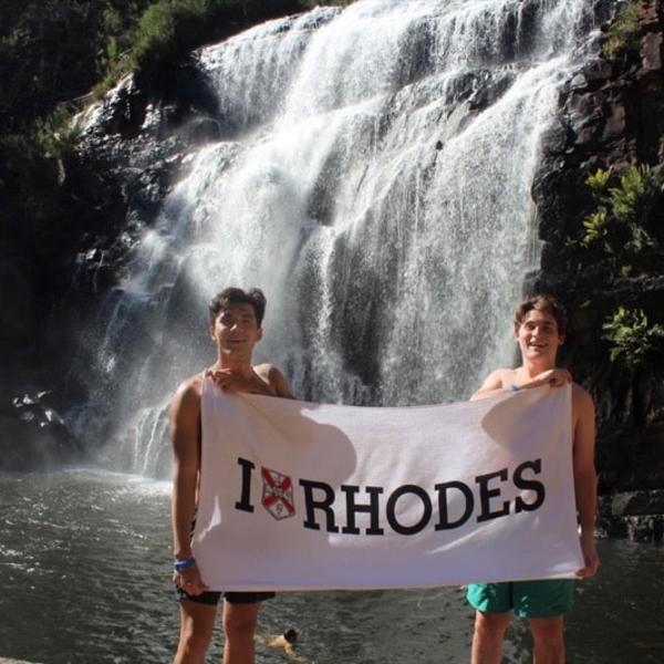 Two young men stand with an "I Heart Rhodes" banner in front of a rushing waterfall.