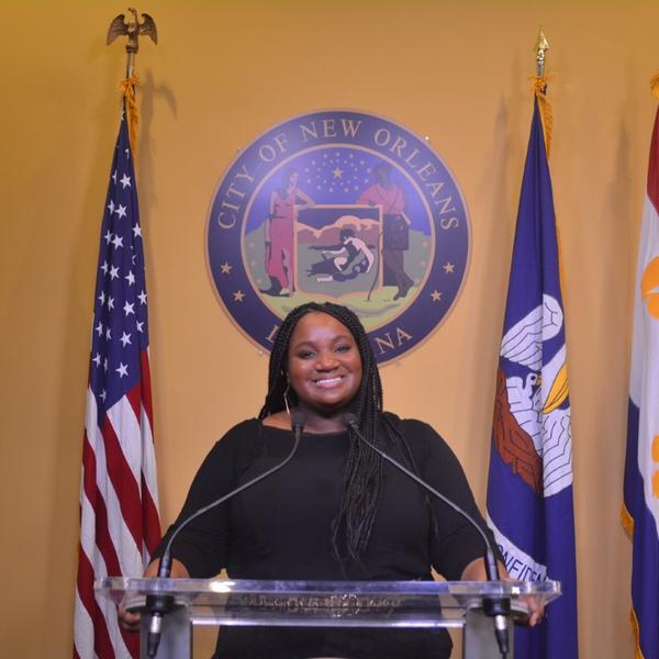 Taylor Jackson stands behind a glass podium in front of the sea of New Orleans and three flags