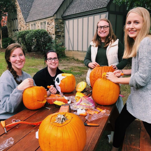Four young women carve pumpkins in foreground of a sorority house