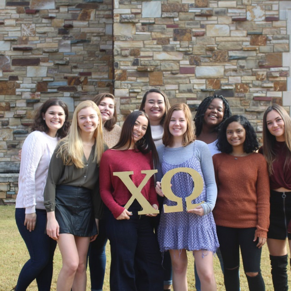 A group of women in foreground of a stone building, holding Chi Omega letters