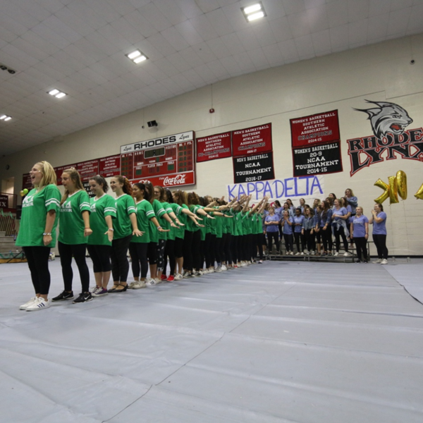 Women in green shirts stand in a dance line in a gym in front of the words Kappa Delta