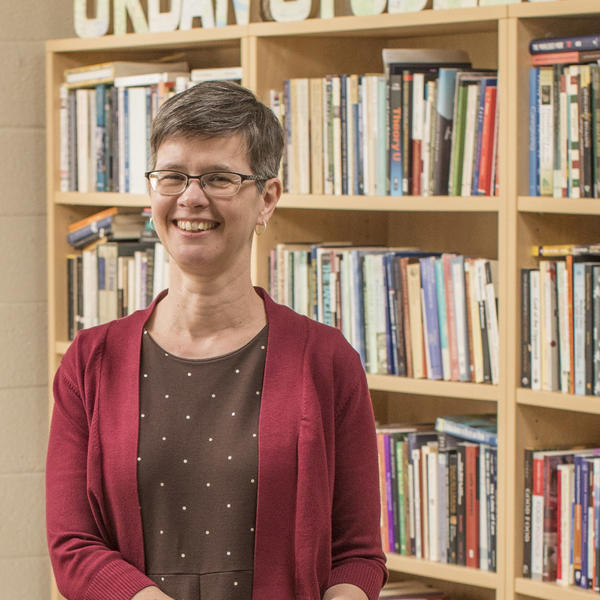 a woman with short hair stands in front of a bookcase with the words Urban Studies on top