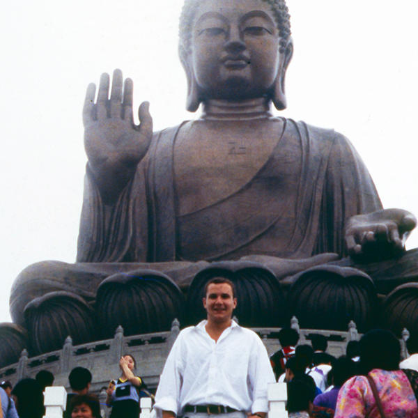 a young man stands in front of a huge statue of the Buddha