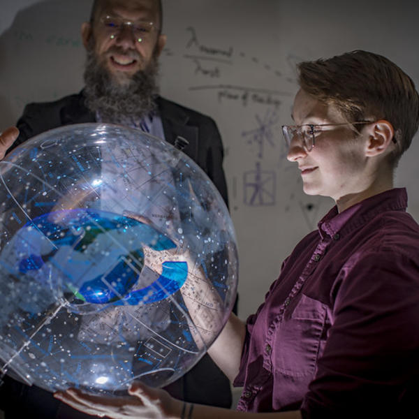 A professor stands behind a clear model of the milky way. Light shines up from beneath the model, onto the face of a student to whom he is explaining it.