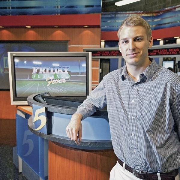 A man in an open-collared button down leans casually against the news desk and WMCTV.