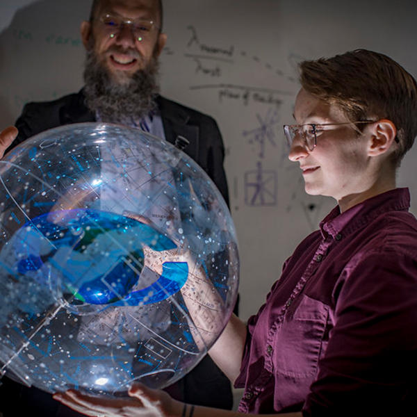 a student holds a globe showing the constellations while a professor looks on