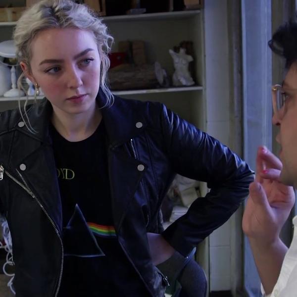 A young woman leans against a work table while talking to her professor.