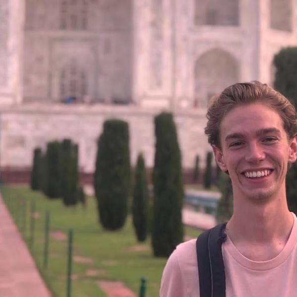 A young man smiles before a formal garden leading to a towering Indian edifice.