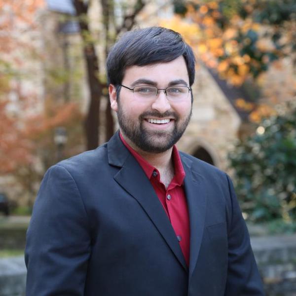 A young, dark haired man smiles into the camera with fall leaves in the background.
