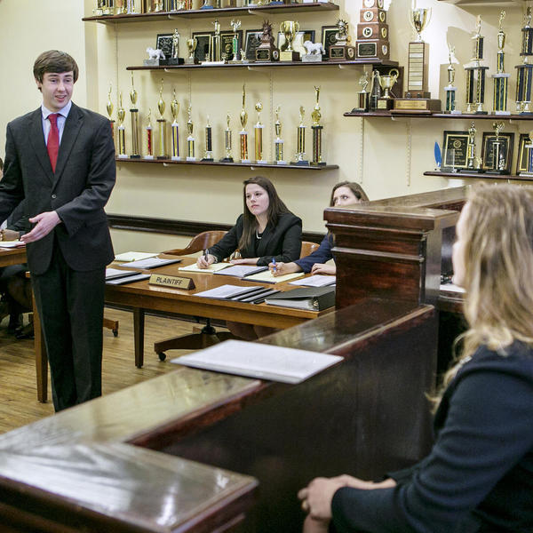 Young man in a suit gestures to evidence as opposing council and a judge look on.