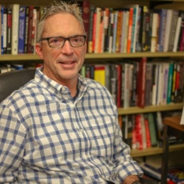 A man in a checked shirt stands in front of a bookshelf.