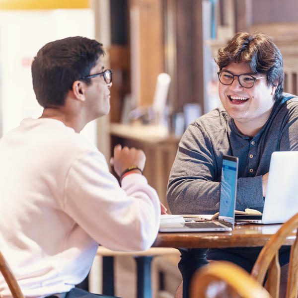 Two male student sit talking at a table. On the table is a laptop with several stickers on the back.