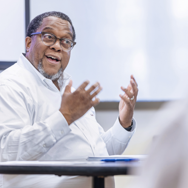 A teacher in a white shirt makes an excellent point while seated in front of a whiteboard.