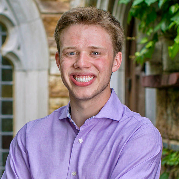 A young man smiles for the camera while standing in front of a stained glass window