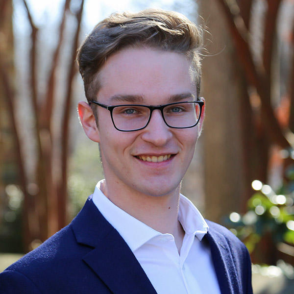 A young man in a blue blazer stands in front of trees.