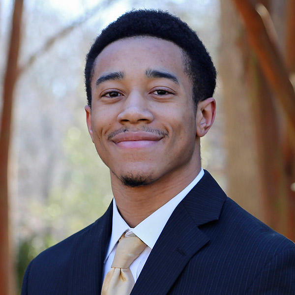 A young man in a suit with a tasteful gold tie stands in front of crepe myrtle trees.