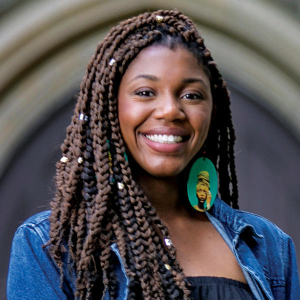 A young woman with dreads stands under a stone arch.