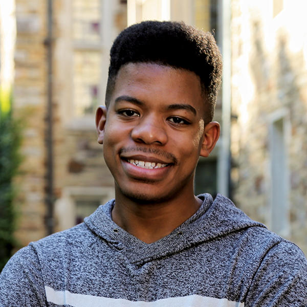 A young man in a grey hoodie stands in front of a stone building.