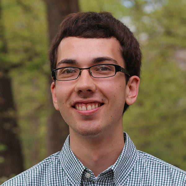A young man with glasses stands smiling in a wooded area.