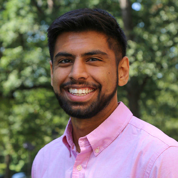 A young man in a pink button down shirt stands smiling in front of bushes.