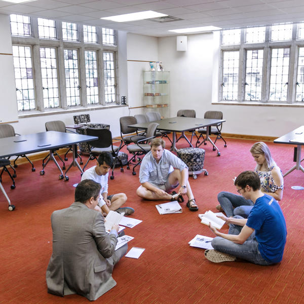 A professor and students sit in a circle on red carpet reviewing for an upcoming class.