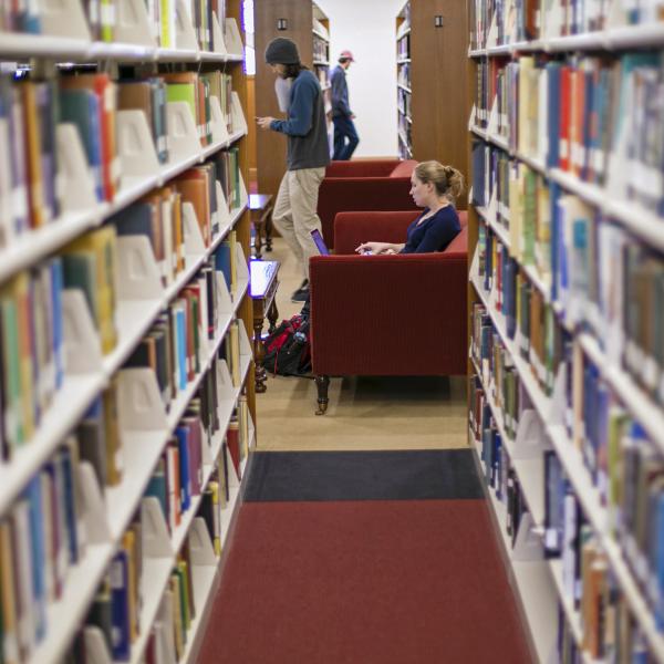 Between two rows of bookshelves, a student sits in a chair and another walks by