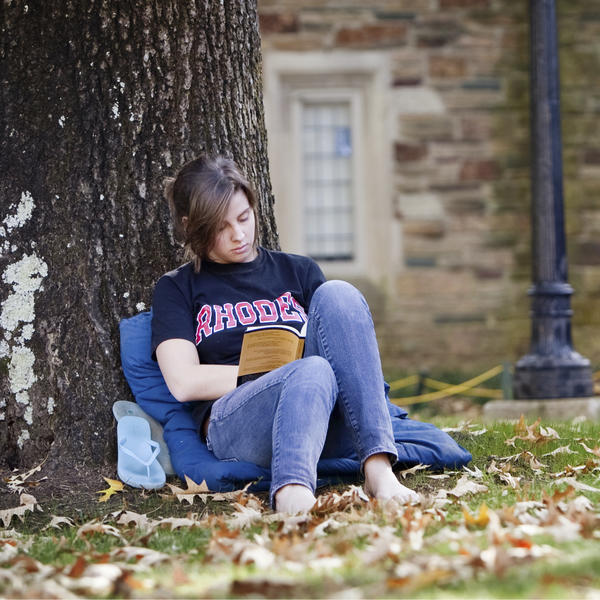 A barefoot young woman in a Rhodes tshirt and jeans sits leaning against a tree reading a book.