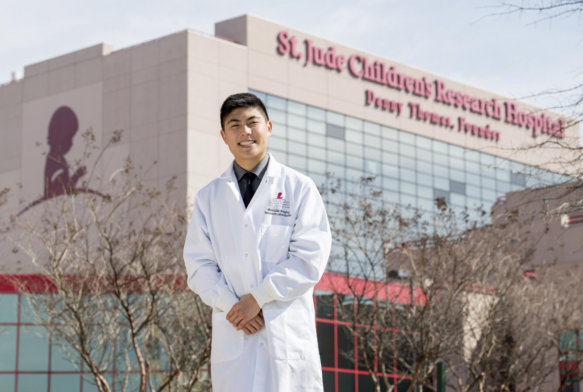 A young man in a white coat stands outside St. Jude Children's Research Hospital.