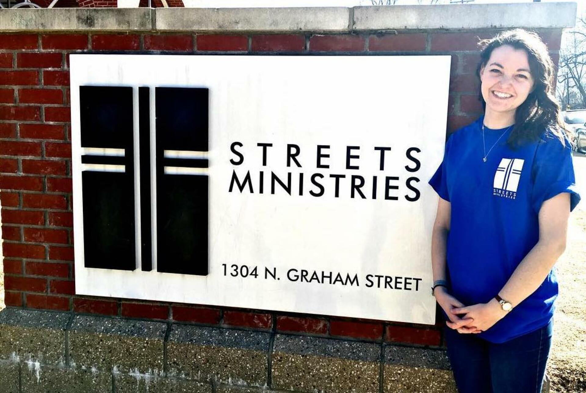 A woman in a blue t-shirt stands in front of a sign inlayed in brick that says Streets Ministries