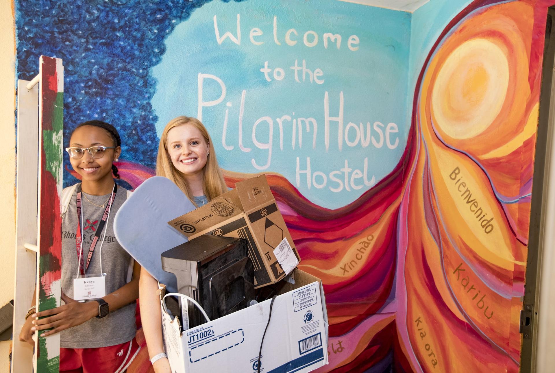 Two students carrying boxes in front of a mural that reads "Pilgrim House Hostel"