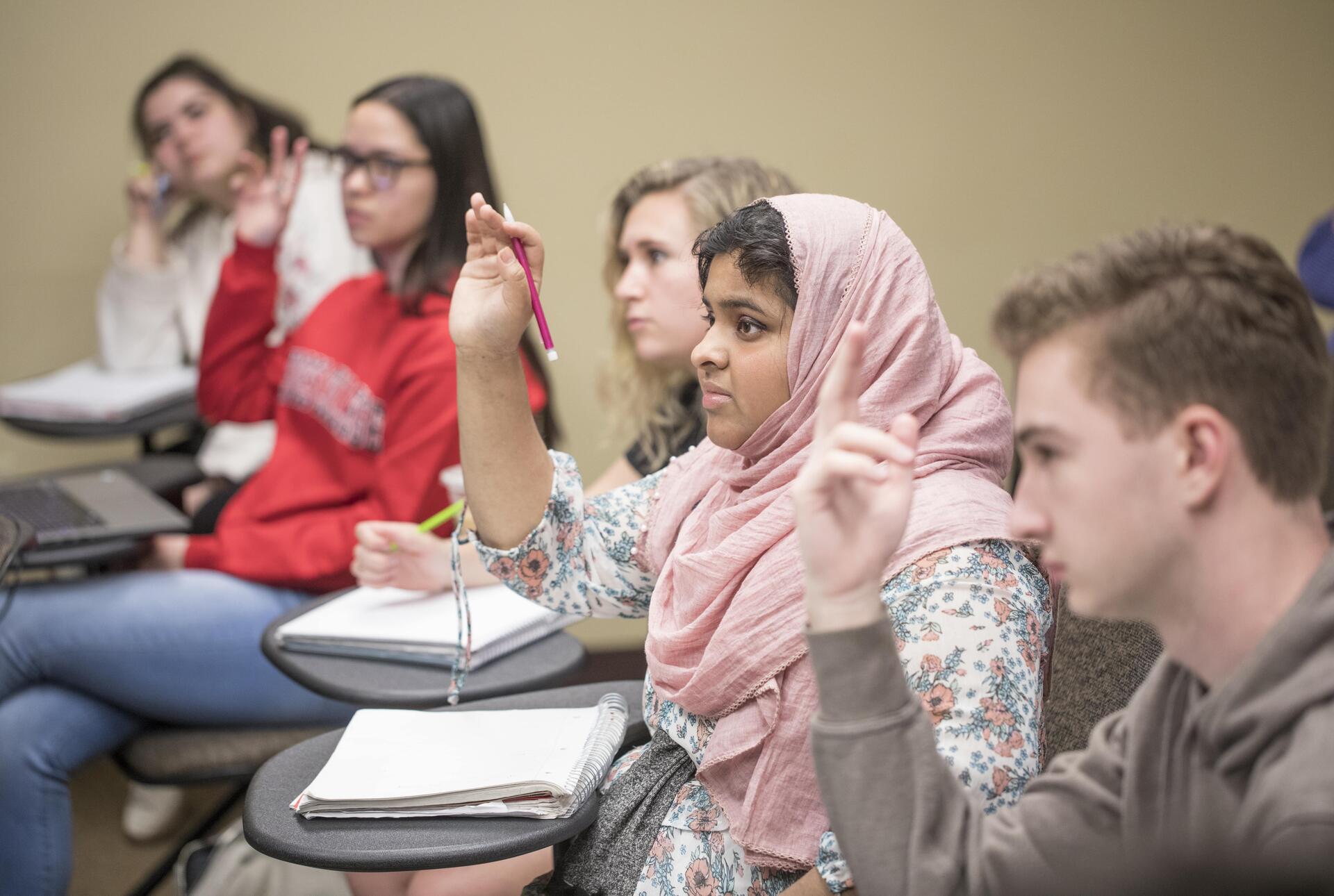 Five students are sitting at their desks, three with hands raised; the focus is on a young woman in a pink hijab