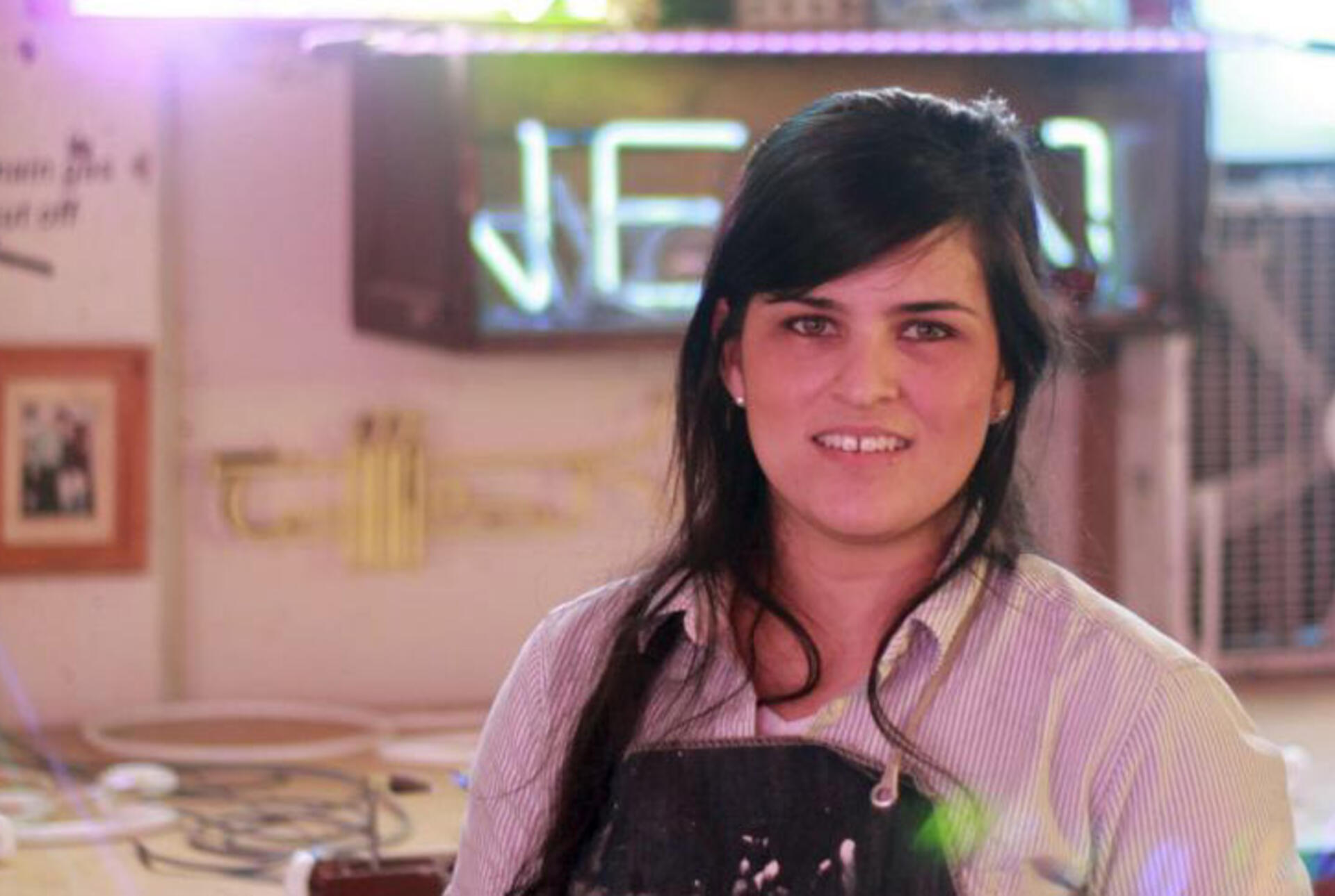 a young woman with dark hair stands in a studio in front of a neon sign