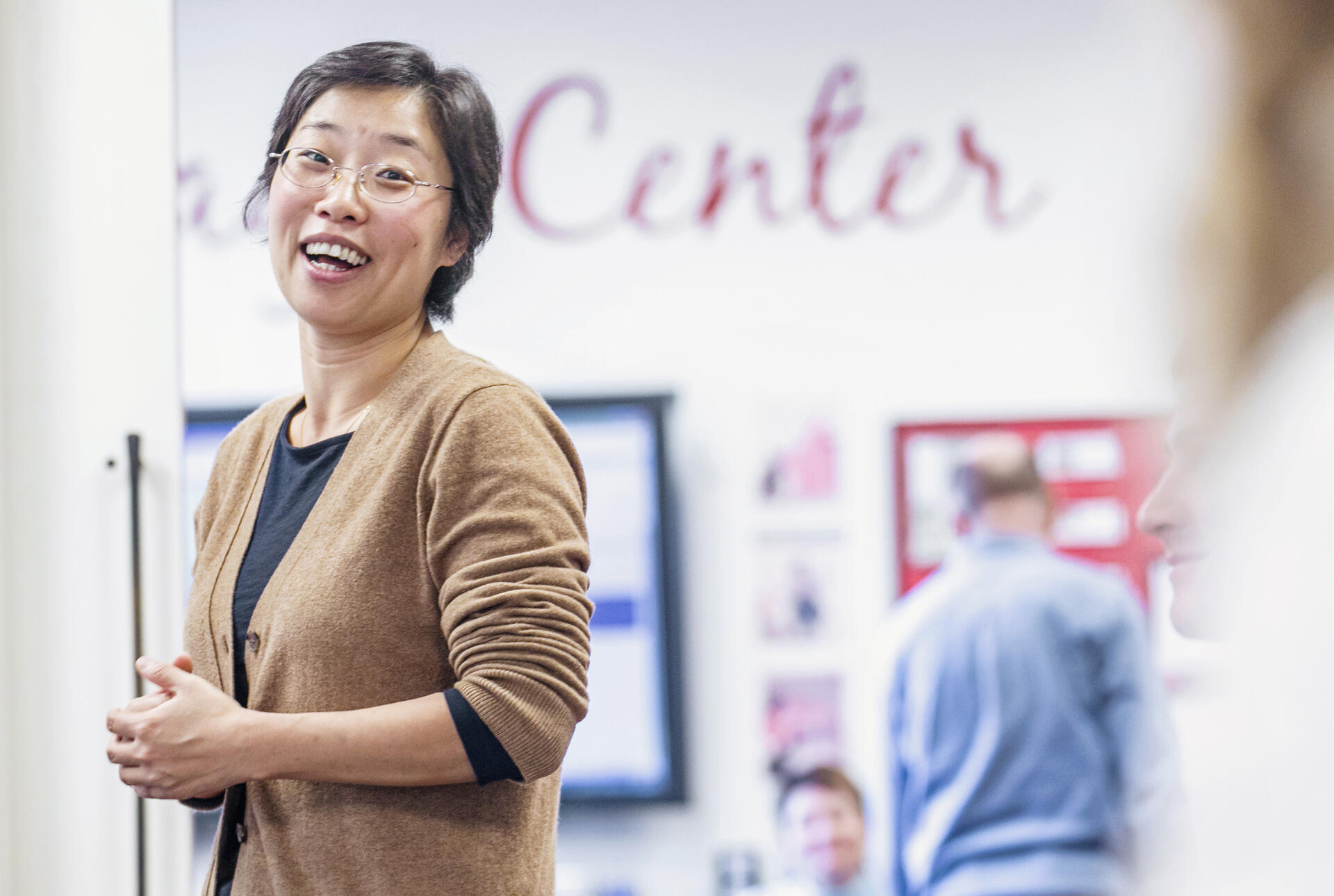 An Asian woman in a tan sweater laughs with students