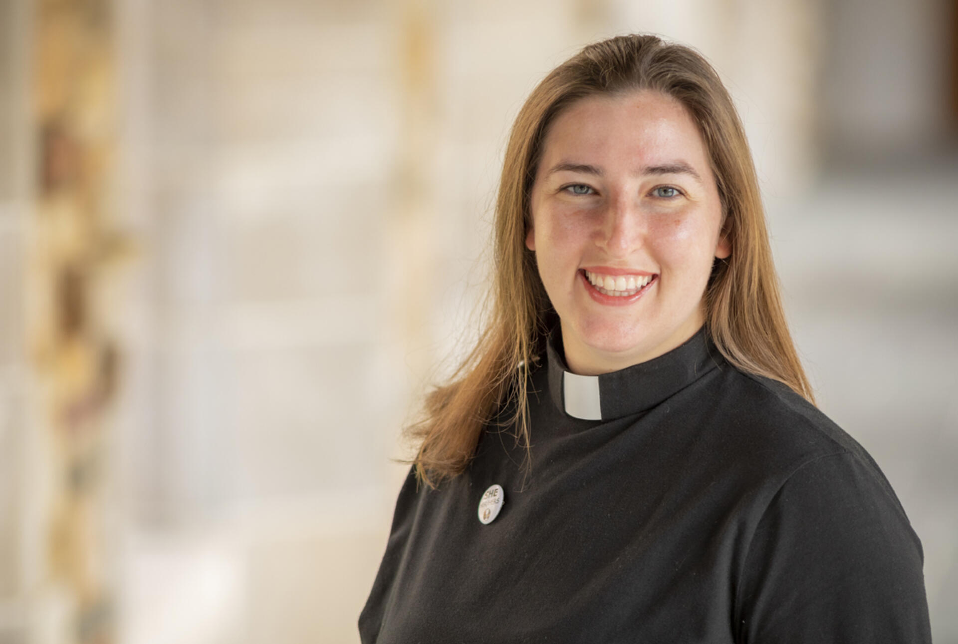 A young, dark haired woman in clerical vestments smiles at the camera
