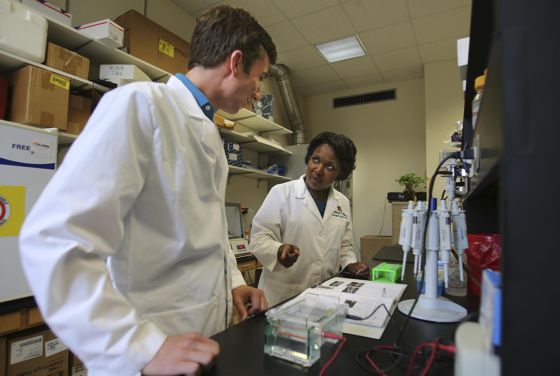 Two researchers in white coats stand at a lab table reviewing notes.