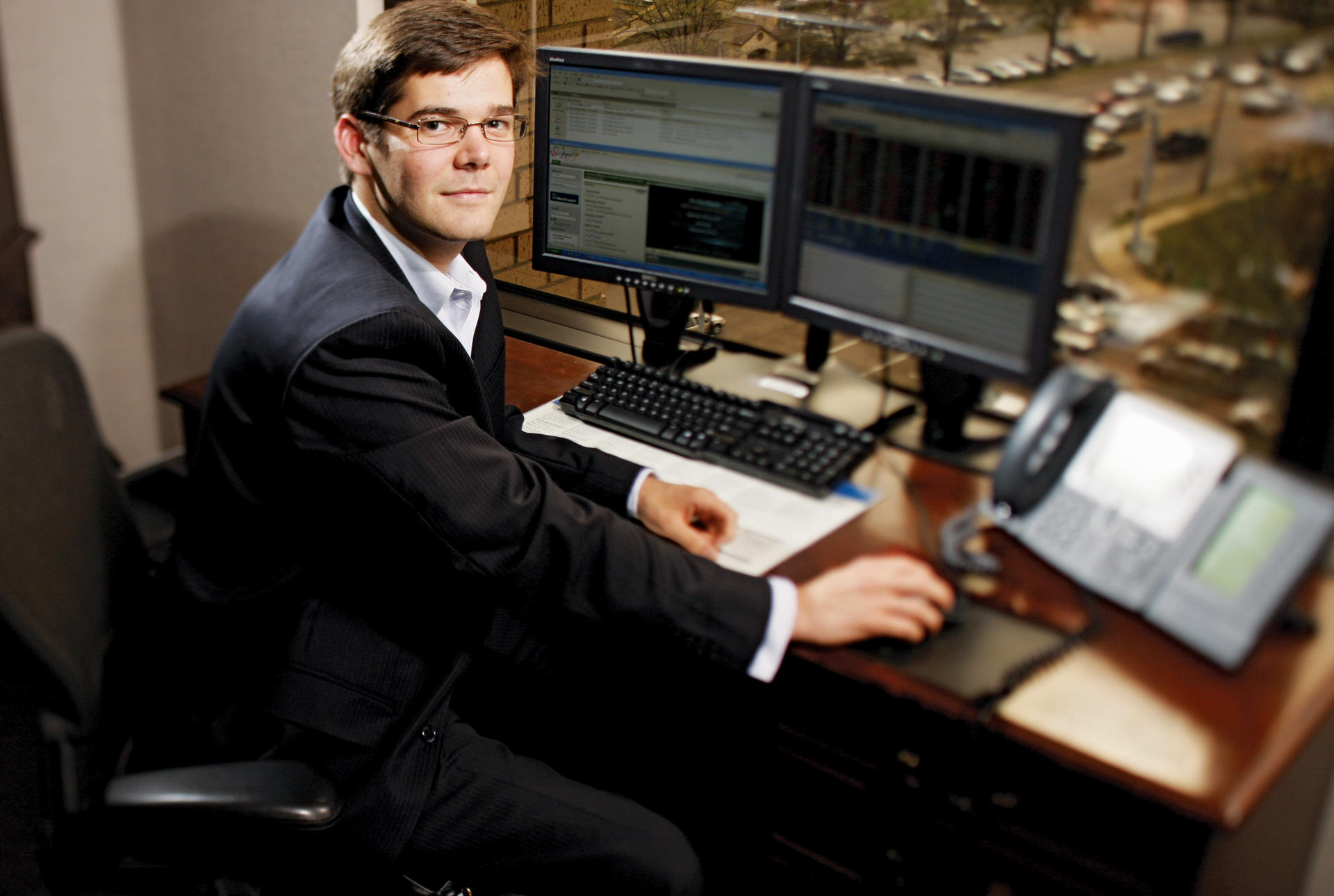 A young man in a suit sits at a computer, looking back at the camera.