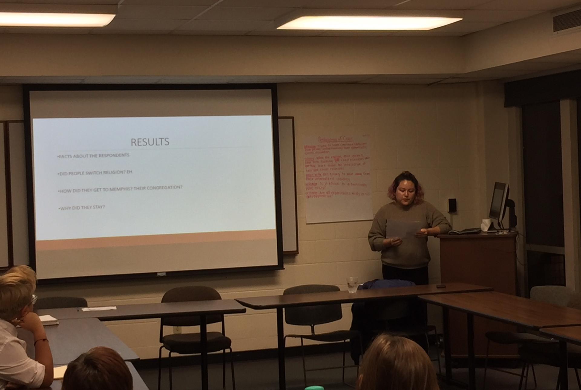 A student stands presenting near a projection screen in a classroom 