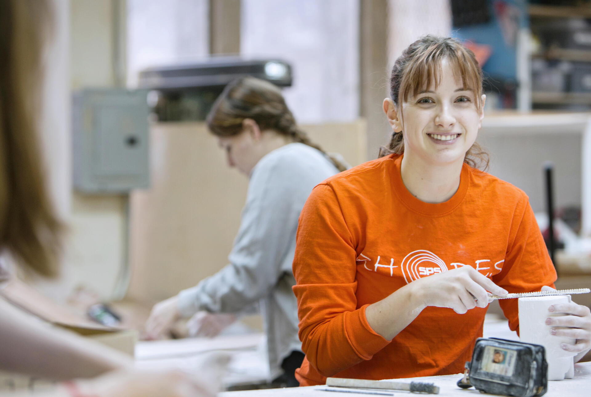 A brown haired young woman in an orange sweater smiles for the camera.