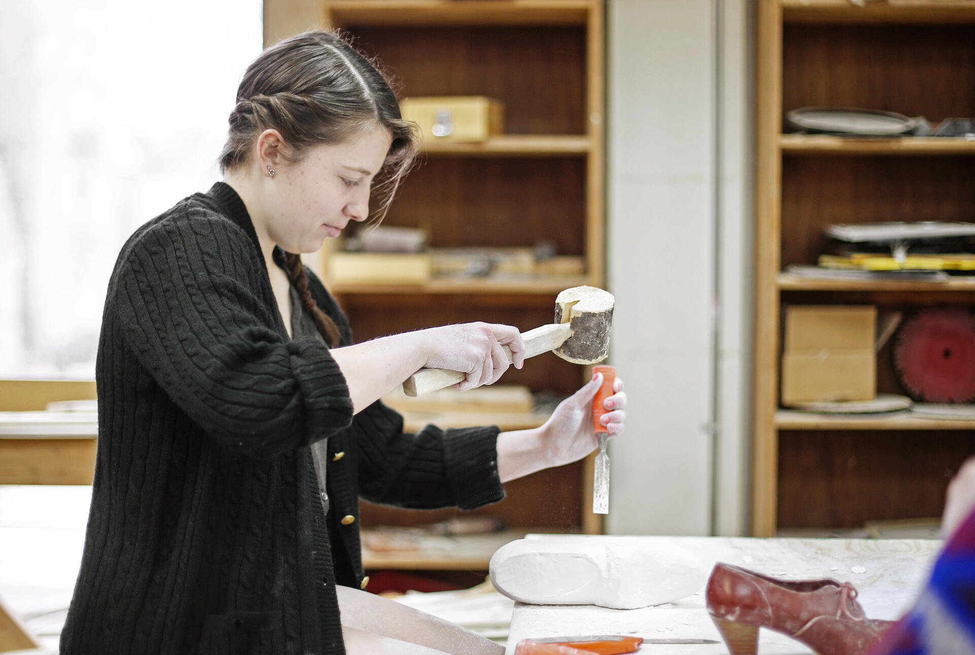 A woman in a dark jacket uses a large wooden mallet and an orange chisel to knock away at a granite carving of a shoe.