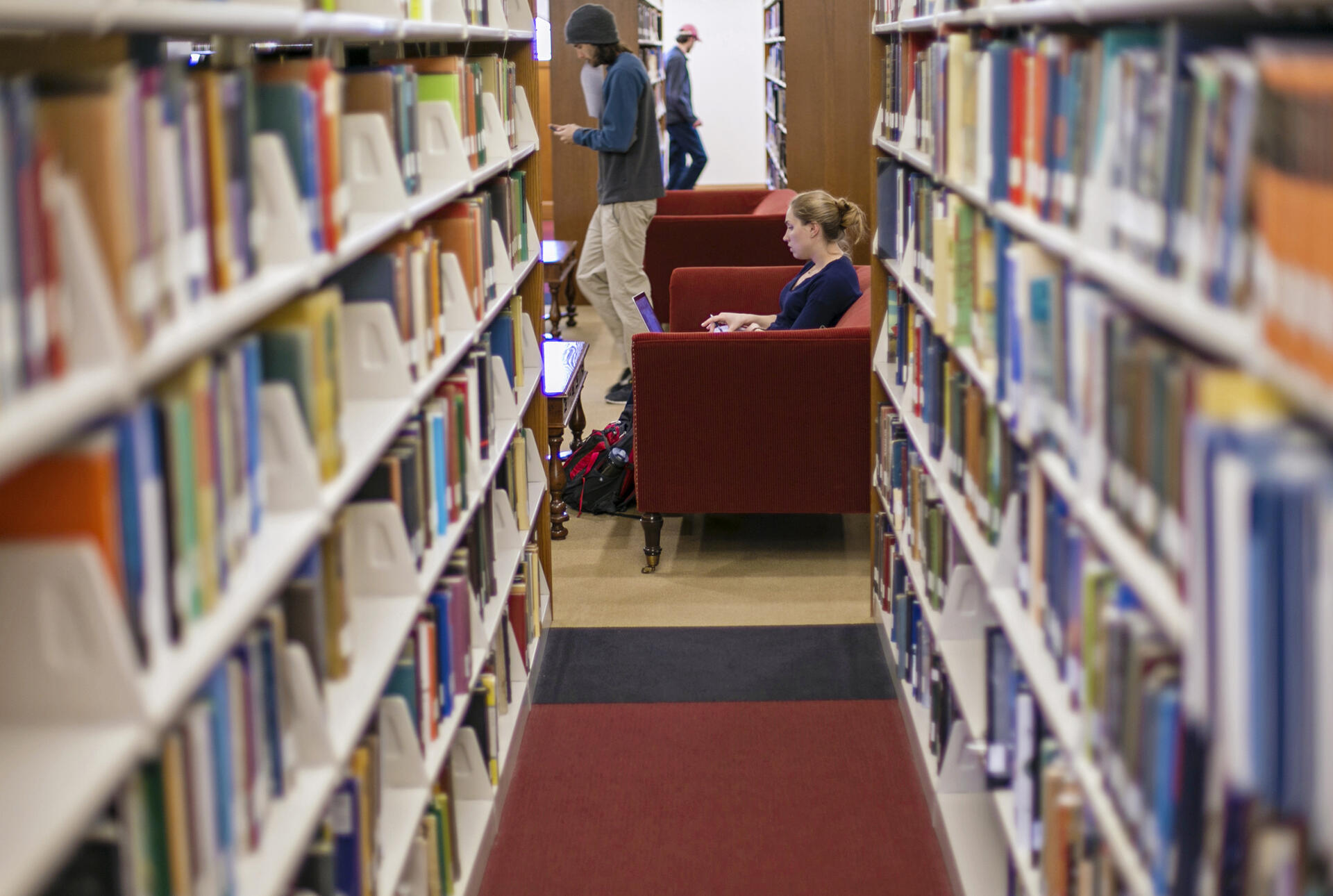 Between two rows of bookshelves, a student sits in a chair and another walks by