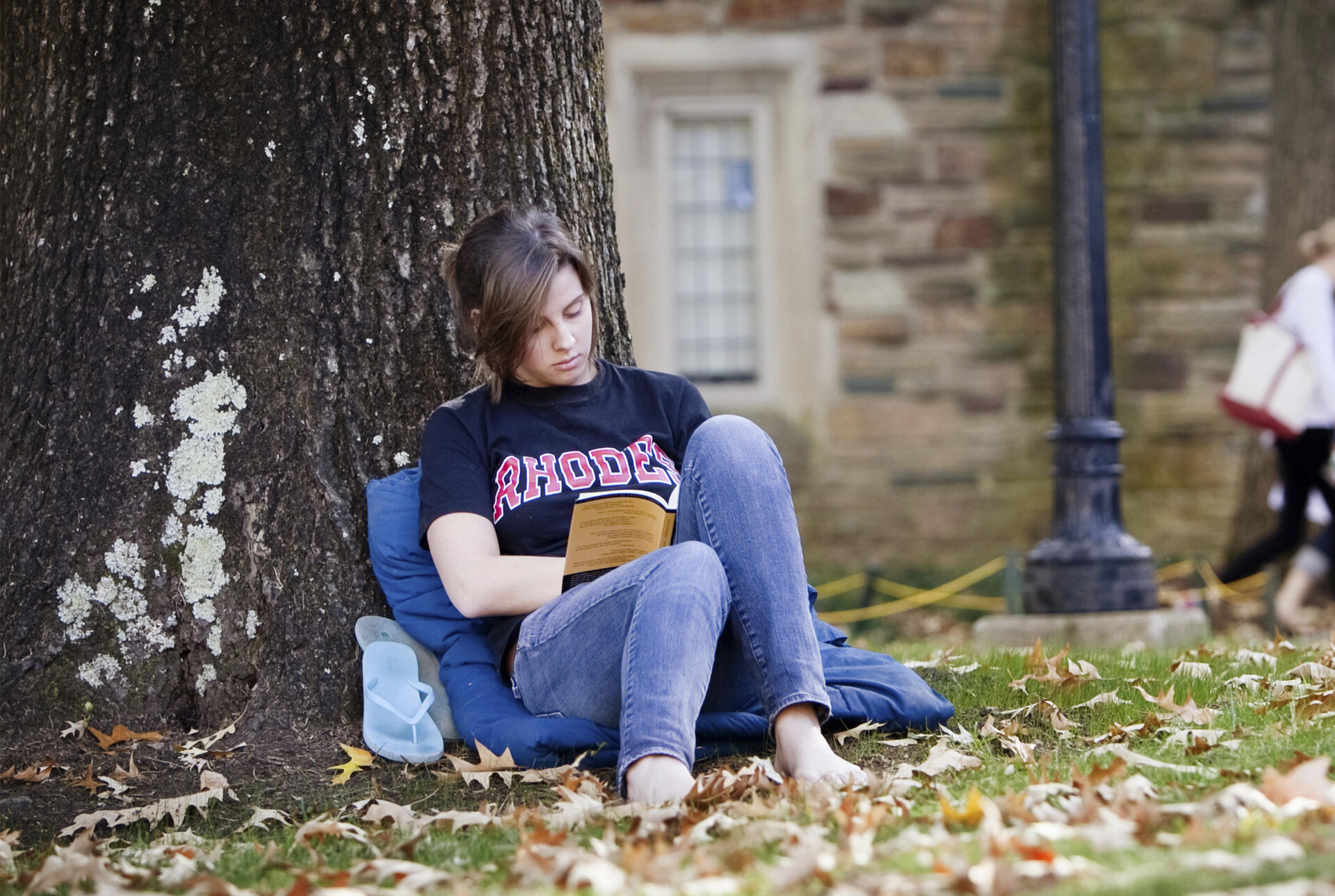 A barefoot young woman in a Rhodes tshirt and jeans sits leaning against a tree reading a book.